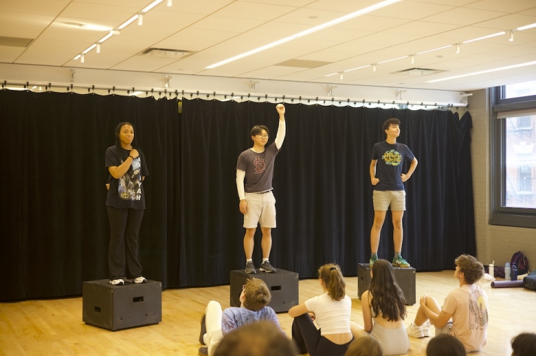 Three Tisch Summer High School Drama students stand on raised boxes on a stage during their final presentations on the last day of class. The student in the middle has an arm raised above his head. The student to the left holds an arm over her heart and the student to the right poses with arms akimbo